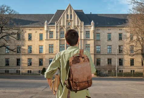 A student with a brown backpack standing in front of a large historic college building