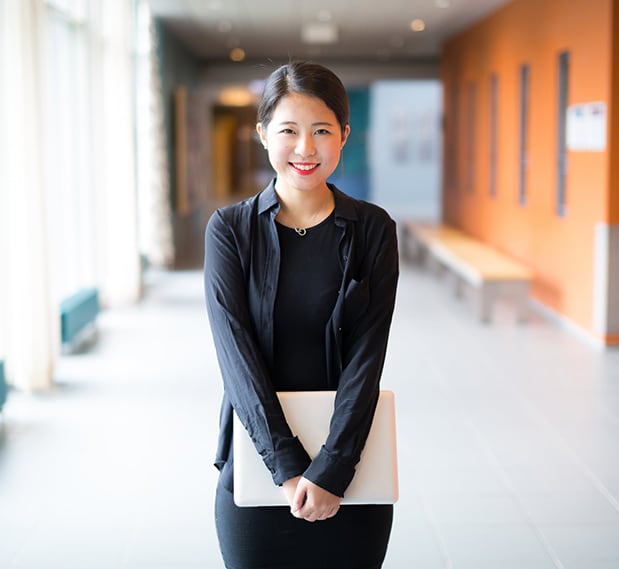 A female student dressed in black holding a laptop and smiling in a school hallway