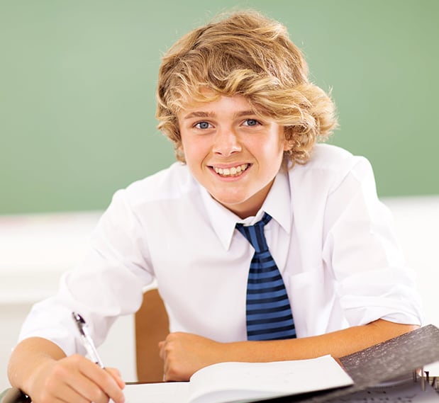 Young male student in a white shirt and striped tie smiling while writing in a notebook