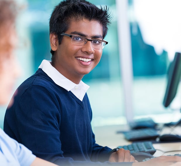 A male student in glasses smiling while seated at a computer in a classroom