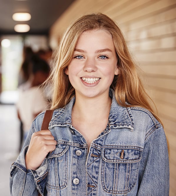 A young high school student smiling while standing in a school hallway with other students behind her