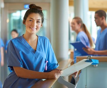 A female medical student smiling in scrubs while standing at a nurse’s station with others in scrubs behind her