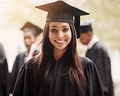 A high school girl graduate in a cap and gown with classmates in background