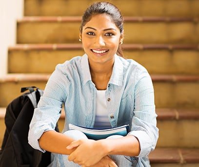 A smiling female high school student of Indian descent sitting on stairs inside school
