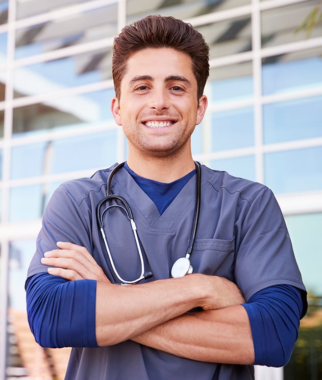 A male medical student in scrubs smiling with arms crossed outside a modern building