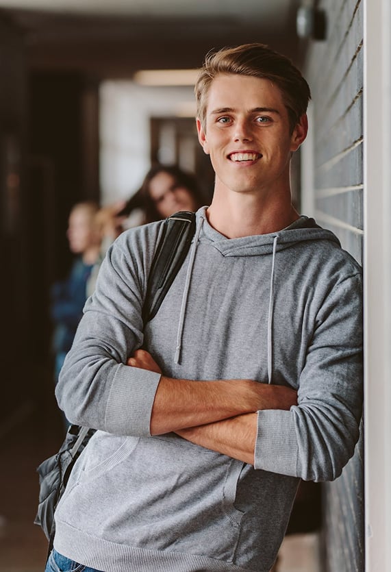 A tall high school boy leaning against hallway wall with arms crossed with other students behind him