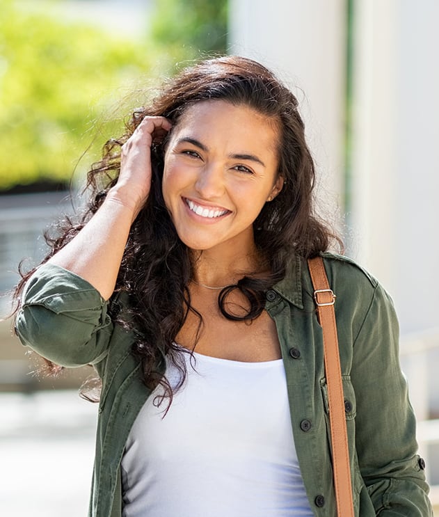 A female student smiling outdoors while holding her hair, wearing a green jacket and white top