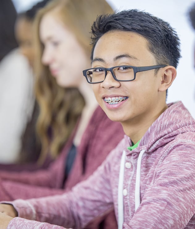 A male student with braces and glasses smiling while sitting in class with other students