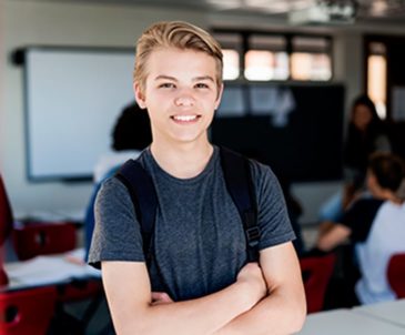 A teenage boy with a backpack smiling while standing confidently in a classroom with desks and students behind him