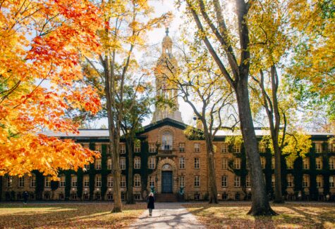 A student walks toward a historic university building surrounded by colorful fall trees