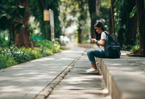 A student with a backpack sitting on a low ledge in a tree-lined walkway, looking at her phone