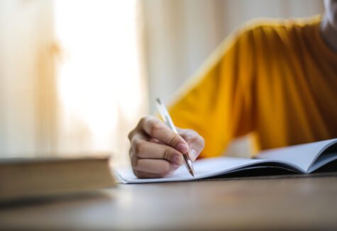 A student in a yellow shirt writing in a notebook at a wooden desk near a window