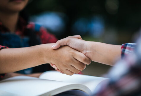 Two students shaking hands over an open book during an outdoor study session