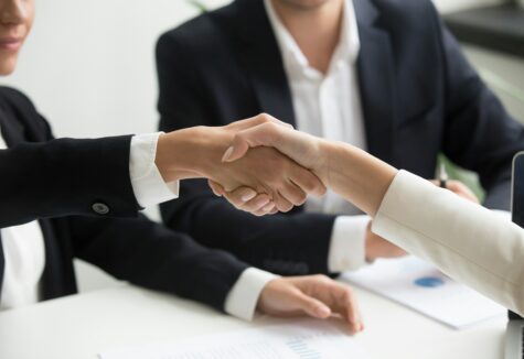 Two professionals shake hands across a desk in a corporate setting, symbolizing agreement, collaboration