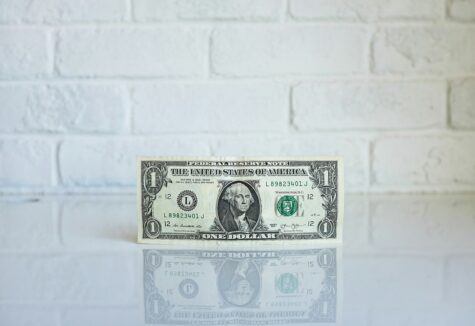 A single U.S. dollar bill standing upright against a white brick wall with a reflective surface