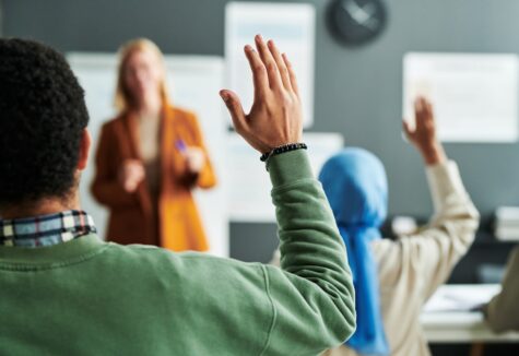 Two students raise their hands to participate while a teacher leads a discussion in front of a classroom