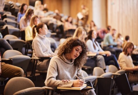Several students sit spaced out in a tiered lecture hall, attentively taking notes during class