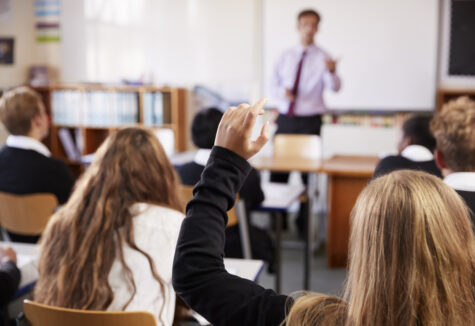 Female student raising their hand in a classroom while a teacher stands at the front