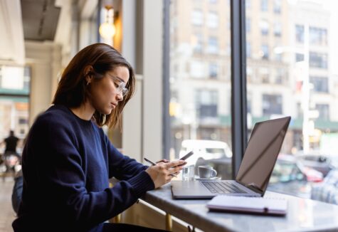 A student in glasses sits by a window in a cafe, working on her phone with a laptop open in front of her