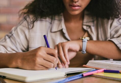 A student sits at a desk, writing in a notebook with colorful pens and study materials spread around