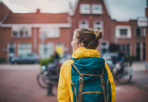 A student wearing a yellow jacket and teal backpack looks toward a campus building with bikes nearby