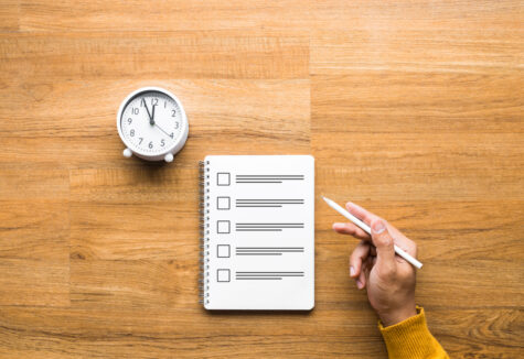 Hand holding a pen above a checklist notebook with a clock on a wooden desk