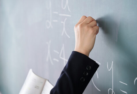 A teacher's hand writing algebraic equations on a green chalkboard while holding an open textbook