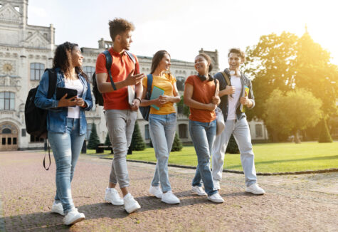 Five students walk together outside a college building, smiling and carrying books