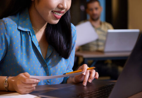 A student smiles while reading documents in front of her laptop during a classroom session with others in the background