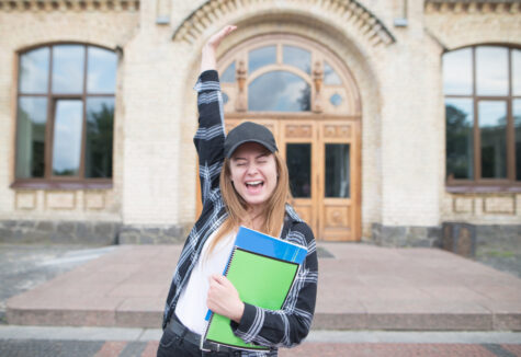 A student raises her arm in celebration while standing outside a university building