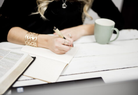 A woman writes in a lined notebook beside an open book and green mug on a white wooden desk