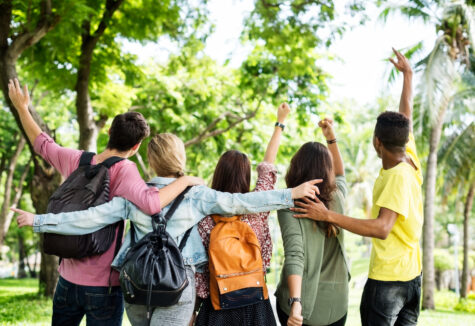Group of five students with backpacks walking through a park with arms raised, seen from behind