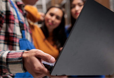 A student holds a laptop while others in the background observe and listen in a casual group setting