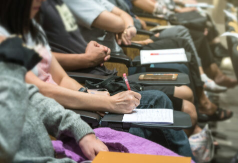 Several students sit in a lecture hall taking handwritten notes during a class session