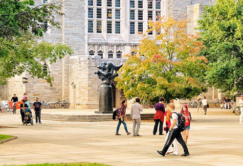 College students walking across a university campus with bikes, trees, and historic buildings in the background