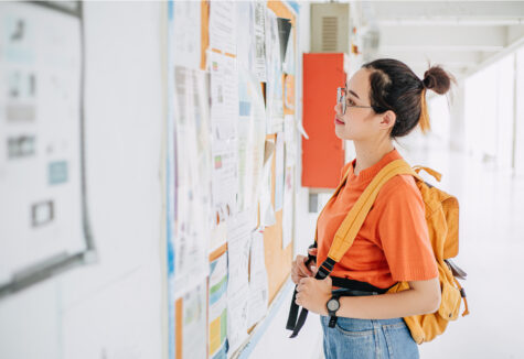 A student in orange top with a yellow backpack reading a bulletin board in a school hallway