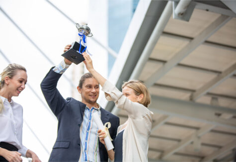 A man holding up a trophy as two women celebrate beside him outside a modern building