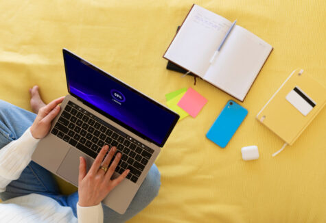 A person using a laptop surrounded by notebooks, phone, and sticky notes on a yellow bedspread