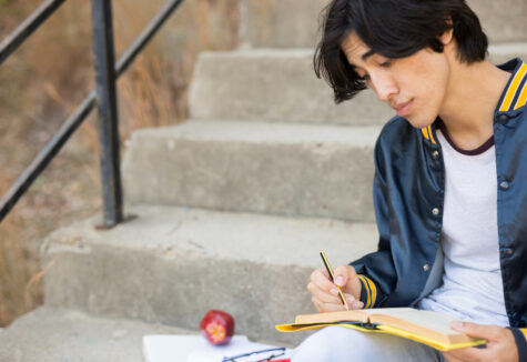 A student sitting on outdoor steps, writing in a notebook with books and an apple beside him