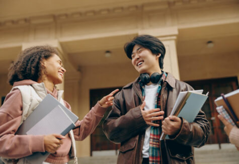 Two college students smiling and talking while holding notebooks outside a campus building