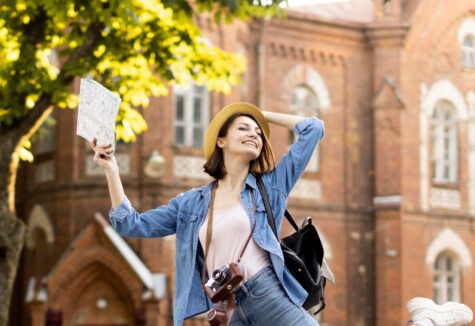 Smiling student holding a map, wearing a hat and backpack in front of a historic college building