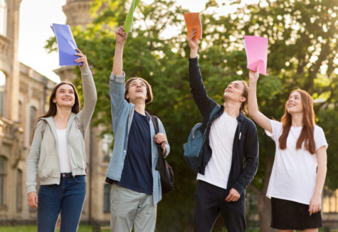 Four smiling students outside holding colorful folders in the air in front of a college building
