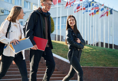 Three students walking down steps outside a university with international flags in the background