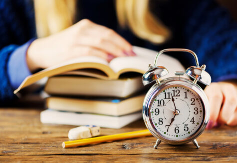 Alarm clock on a wooden table in front of stacked books and a person studying