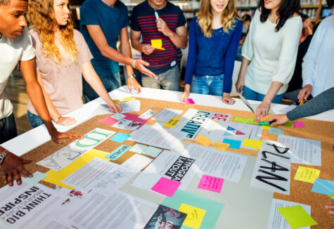 Group of students gathered around a large table filled with notes, charts, and colorful post-its, actively organizing ideas for a project