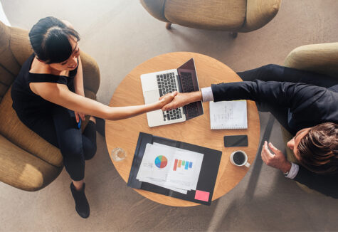 Two professionals shake hands at a round table with charts and a laptop