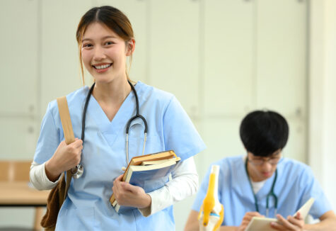 A smiling nursing student holds books and a stethoscope in a classroom
