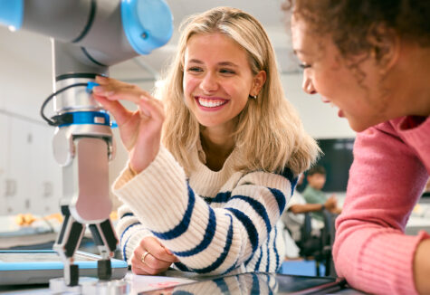 Two female students smile while working with a robotic arm in a classroom
