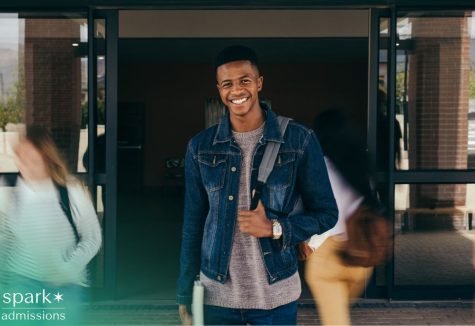 Smiling college student with a backpack stands outside a campus building entrance, holding a notebook as other students walk by.